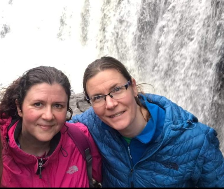 Picture of dark haired woman in pink jacket and woman with glasses in blue jacket in front of waterfall